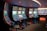 A row of brightly lit slot machines in a casino setting, each with colorful displays and various game themes. The machines are lined up against a dark wall and are unoccupied, with cushioned chairs placed in front of them. The area is dimly lit except for the glow from the screens, creating a vibrant and engaging atmosphere.