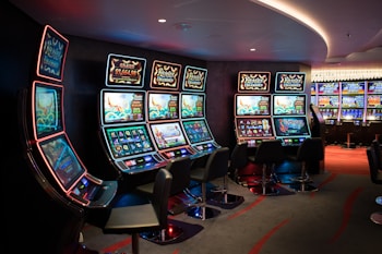 A row of brightly lit slot machines in a casino setting, each with colorful displays and various game themes. The machines are lined up against a dark wall and are unoccupied, with cushioned chairs placed in front of them. The area is dimly lit except for the glow from the screens, creating a vibrant and engaging atmosphere.
