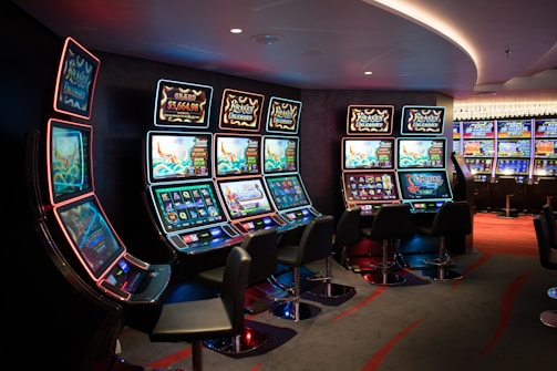A row of brightly lit slot machines in a casino setting, each with colorful displays and various game themes. The machines are lined up against a dark wall and are unoccupied, with cushioned chairs placed in front of them. The area is dimly lit except for the glow from the screens, creating a vibrant and engaging atmosphere.