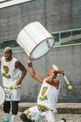 Two men are engaged in a lively performance, wearing matching outfits with colorful patterns. One man is energetically playing a large, white drum while the other is raising a stick with a vibrant tassel. The background consists of a blue tiled wall, adding a dynamic urban setting to the scene.