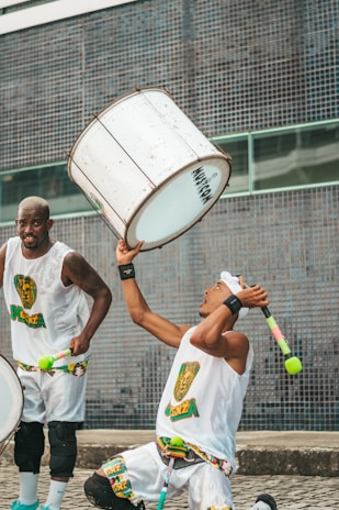 Two men are engaged in a lively performance, wearing matching outfits with colorful patterns. One man is energetically playing a large, white drum while the other is raising a stick with a vibrant tassel. The background consists of a blue tiled wall, adding a dynamic urban setting to the scene.