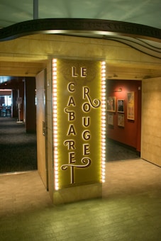 A warmly lit entrance to a venue with an illuminated sign displaying 'Le Cabaret Rouge'. The sign features elegant, vintage-style lettering bordered by rows of lightbulbs. The background shows a hallway with framed artwork on red walls, creating an inviting atmosphere.