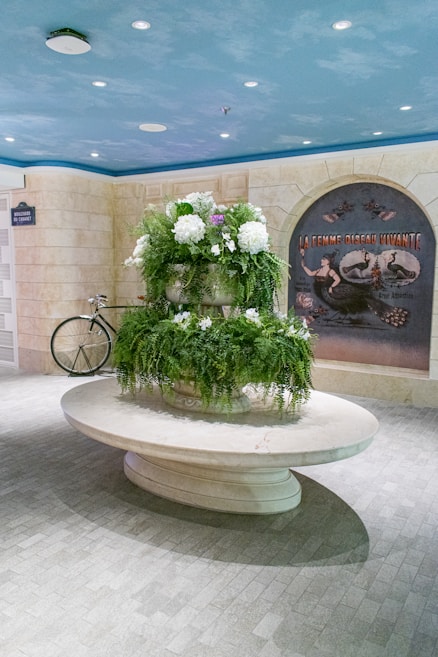 A decorative indoor setting features a large ornate planter on an oval marble table, filled with lush green ferns and white flowers. A vintage-style poster is mounted on a stone-textured wall, depicting an old-time attraction with a mermaid-like figure. Blue ceiling with spotlights creates a cool ambiance. A bicycle is casually leaned against the wall in the background.