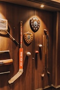 A variety of sports memorabilia displayed on a wooden wall, including a hockey stick, cricket bat, golf clubs, plaques, and balls. The lighting casts warm tones and highlights the vintage and rustic feel of the items.