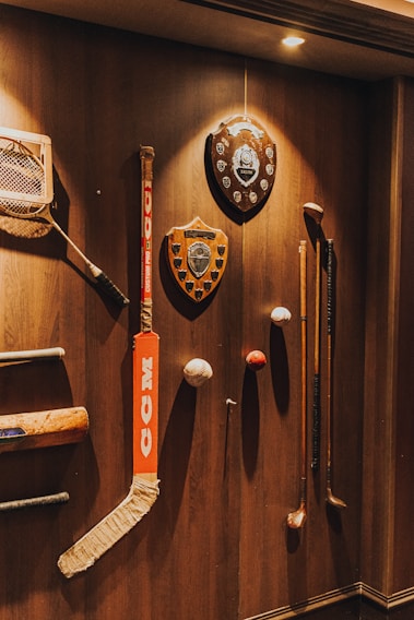 A variety of sports memorabilia displayed on a wooden wall, including a hockey stick, cricket bat, golf clubs, plaques, and balls. The lighting casts warm tones and highlights the vintage and rustic feel of the items.