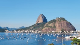 A large bay filled with numerous sailboats and a sprawling cityscape at the base of large, rocky hills. The hills, covered in greenery, rise dramatically with a clear blue sky overhead.