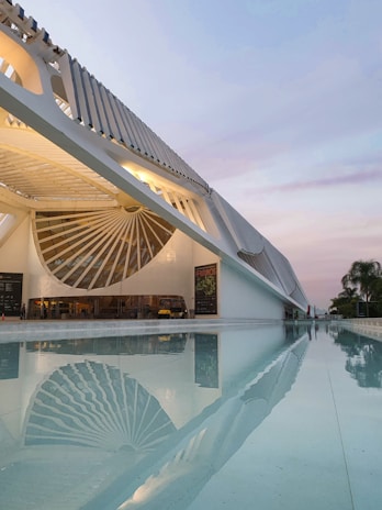 A striking modern architectural structure with a large, well-lit facade is reflected beautifully in a serene body of water. The design features bold geometric shapes and lines, with a blue and purple sky in the background, adding to the overall aesthetic.