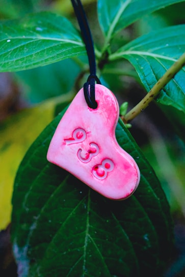 A heart-shaped pink charm with the number 938 is hanging from a black string against a backdrop of lush green leaves.