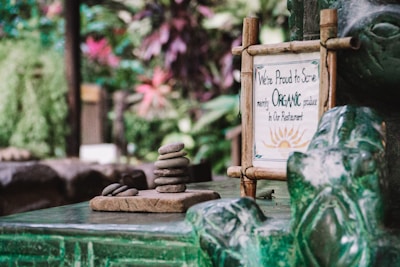 In a lush, green setting, a small sign framed in a rustic wooden frame indicates a commitment to serving organic produce. Next to the sign, there is a small stack of smooth stones placed neatly on a stone platform, giving a sense of tranquility and balance.