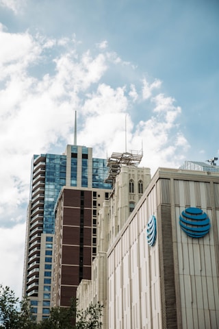 Several modern skyscrapers stand tall against a partly cloudy sky. The buildings have varied architectural styles, with glass and stone facades. A prominent logo with concentric circles is visible on one of the buildings.