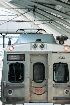 A front view of an RTD train at a station, displaying its metallic surface with visible headlights and a digital sign indicating destination 'Union Station'. The scene includes the overhead wires and part of the station structure.