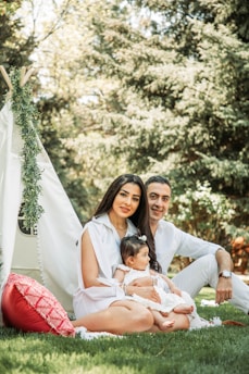 A family enjoying a day outdoors with a teepee tent set up on the grass. A woman with long dark hair and a man, both dressed in light-colored clothing, sit on the ground. The woman is holding a baby who is also wearing light clothes. The scene is surrounded by lush green trees, and there's a red cushion next to the tent.