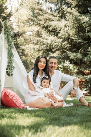 A family of three is sitting on a blanket in a grassy area with trees in the background. They are dressed in white and seated in front of a decorative teepee adorned with greenery. A red pillow is placed next to them.