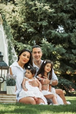 A family of four is sitting on the grass in an outdoor setting, surrounded by trees. The family includes a man, a woman, and two young children, all dressed in white. There is a decorative tent with greenery and a lantern nearby.
