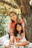 A smiling mother and teen sitting together outdoors, sharing a relaxed moment with warm sunlight filtering through trees.