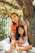 A smiling mother and teen sitting together outdoors, sharing a relaxed moment with warm sunlight filtering through trees.