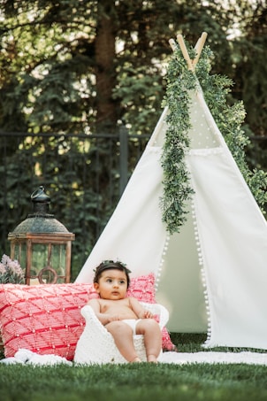 A baby wearing a floral headband sits comfortably on a knitted cushion, surrounded by a white teepee adorned with greenery. In the background, a decorative lantern and vibrant red pillow add to the whimsical outdoor setting. Lush green grass and trees contribute to the natural and serene environment.