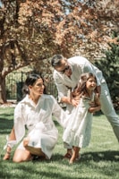 A family of three is enjoying time together outdoors on a sunny day. The young girl is wearing a white dress and is being supported by her father, who is bending over and smiling. The mother, also dressed in white, is kneeling on the grass and looking at her daughter affectionately. The background features lush green grass and a tree with brown and green leaves, suggesting a warm, pleasant atmosphere.