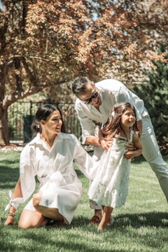 A family of three is enjoying time together outdoors on a sunny day. The young girl is wearing a white dress and is being supported by her father, who is bending over and smiling. The mother, also dressed in white, is kneeling on the grass and looking at her daughter affectionately. The background features lush green grass and a tree with brown and green leaves, suggesting a warm, pleasant atmosphere.