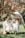 A family of three is enjoying time together outdoors on a sunny day. The young girl is wearing a white dress and is being supported by her father, who is bending over and smiling. The mother, also dressed in white, is kneeling on the grass and looking at her daughter affectionately. The background features lush green grass and a tree with brown and green leaves, suggesting a warm, pleasant atmosphere.