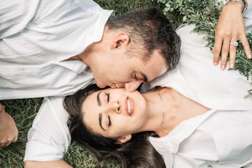 A couple lies on the grass, with the man leaning over to kiss the woman's forehead. They both wear white shirts and are surrounded by greenery. The woman has her eyes closed and smiles softly, indicating a moment of tenderness and affection.