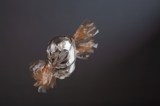 A single piece of candy wrapped in shiny, crinkled foil is placed on a reflective, dark surface. The wrapper extends out to the sides with a clear, cellophane-like material.