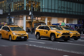 Three yellow taxis are driving on a city street in front of a glass storefront building. Pedestrians are visible on the sidewalk, and the building features a sign for a clothing store.