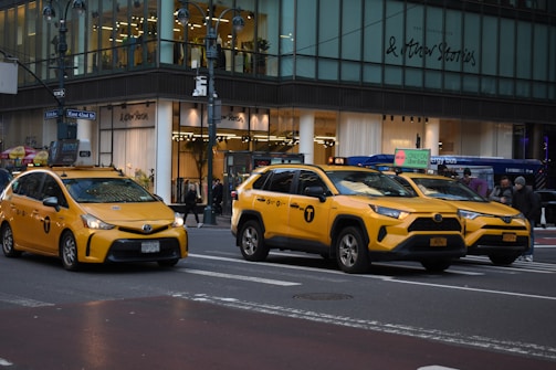 Three yellow taxis are driving on a city street in front of a glass storefront building. Pedestrians are visible on the sidewalk, and the building features a sign for a clothing store.