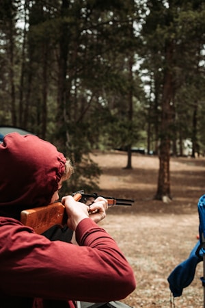 A person wearing a red hoodie is aiming a rifle in an outdoor wooded area. The ground is covered with dry leaves and several trees surround the scene, creating a dense forest backdrop. A blue object is partially visible on the right side of the image.