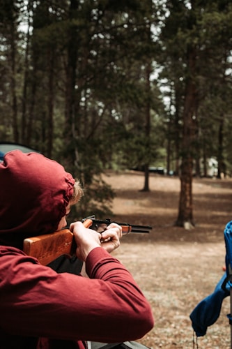 A person wearing a red hoodie is aiming a rifle in an outdoor wooded area. The ground is covered with dry leaves and several trees surround the scene, creating a dense forest backdrop. A blue object is partially visible on the right side of the image.
