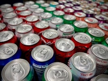 A collection of colorful soda cans is closely packed together, showcasing a variety of brands and flavors. The tops of the cans are prominently displayed, with silver pull tabs visible. The colors range from red, green, blue, to silver, indicating a diverse selection. Some cans display nutrition facts labels, hinting at the variety of options available.