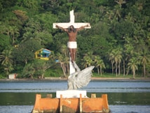 A large statue of Christ on a cross stands amidst water, with lush green palm trees and dense forest in the background. In front of the cross, there's a sculpture of an angel. A small colorful building is visible among the greenery.