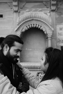 A man and a woman engage in an intimate conversation, standing closely with the woman's hands resting on the man's shoulders. The background features intricate stone architecture, indicative of a historical or cultural site.