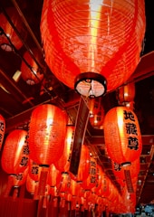 A series of vibrant red paper lanterns with black characters hang closely together, creating a warm and festive atmosphere. The lanterns are intricately decorated and illuminated from within, casting a soft glow.