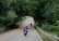Two motorcyclists travel down a winding road lined with lush, green trees, creating a serene and adventurous scene. The leading rider carries a large red backpack. The scene is peaceful with a car visible ahead, suggesting a journey through a rural or forested area.
