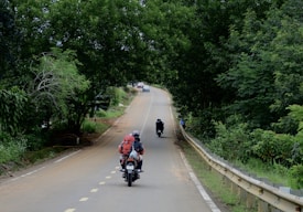 Two motorcyclists travel down a winding road lined with lush, green trees, creating a serene and adventurous scene. The leading rider carries a large red backpack. The scene is peaceful with a car visible ahead, suggesting a journey through a rural or forested area.