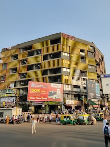 A busy urban street scene with a large, multistory building featuring yellow and black vertical stripes. The building has multiple billboards, including one advertising a 50% discount on bus tickets. Several auto-rickshaws and people are present on the street, depicting a vibrant city atmosphere.