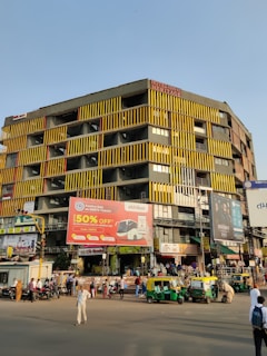 A busy urban street scene with a large, multistory building featuring yellow and black vertical stripes. The building has multiple billboards, including one advertising a 50% discount on bus tickets. Several auto-rickshaws and people are present on the street, depicting a vibrant city atmosphere.
