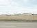 An airport area is visible behind a chain-link fence topped with barbed wire. In the background, there are large buildings, including one with 'Air Canada' signage. A fuel truck with a 'Shell' logo is parked on the grass field. The runway area and airport infrastructure are visible under a cloudy sky.