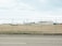 An airport area is visible behind a chain-link fence topped with barbed wire. In the background, there are large buildings, including one with 'Air Canada' signage. A fuel truck with a 'Shell' logo is parked on the grass field. The runway area and airport infrastructure are visible under a cloudy sky.