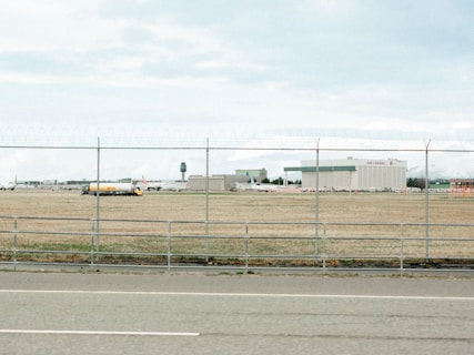 An airport area is visible behind a chain-link fence topped with barbed wire. In the background, there are large buildings, including one with 'Air Canada' signage. A fuel truck with a 'Shell' logo is parked on the grass field. The runway area and airport infrastructure are visible under a cloudy sky.