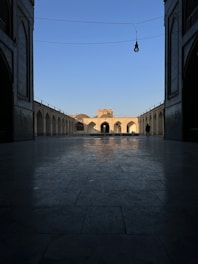 An architectural view captures a courtyard with symmetrical arches and a central building illuminated by sunlight. The scene is framed by a shaded archway in the foreground, enhancing the depth perception. A single figure stands in the distance, adding scale to the composition.