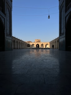 An architectural view captures a courtyard with symmetrical arches and a central building illuminated by sunlight. The scene is framed by a shaded archway in the foreground, enhancing the depth perception. A single figure stands in the distance, adding scale to the composition.
