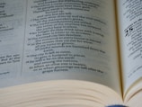 A close-up view of an open book with text on both visible pages. The focus is on one page with poetic or biblical verses written in a column format.