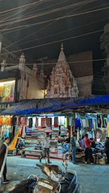 A vibrant street scene featuring a bustling market with various fabrics and clothes hanging from a shop. Several people, including vendors and customers, are visible engaging in activities like browsing and chatting. Overhead wires crisscross the sky, and in the background, a beautifully detailed, illuminated temple stands out against the night sky with ornate architecture.