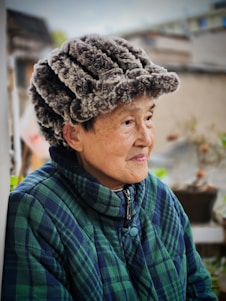 An elderly person wearing a fur hat and a plaid jacket is looking to the side. The background is blurred but suggests an outdoor setting with plants and buildings.