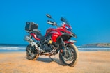 A red adventure motorcycle is parked on a sandy beach with a clear blue sky in the background. The motorcycle is equipped with a top box, and the beach and ocean can be seen extending into the distance with gentle waves lapping against the shore.