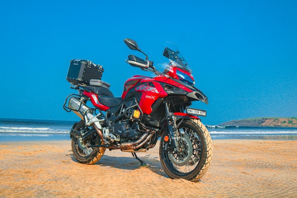A red adventure motorcycle is parked on a sandy beach with a clear blue sky in the background. The motorcycle is equipped with a top box, and the beach and ocean can be seen extending into the distance with gentle waves lapping against the shore.