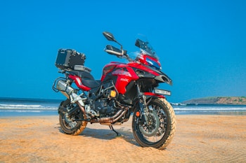 A red adventure motorcycle is parked on a sandy beach with a clear blue sky in the background. The motorcycle is equipped with a top box, and the beach and ocean can be seen extending into the distance with gentle waves lapping against the shore.