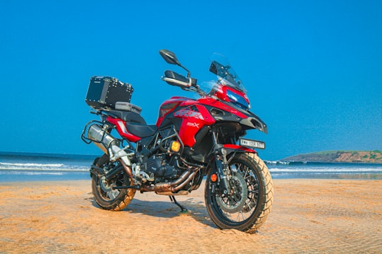 A red adventure motorcycle is parked on a sandy beach with a clear blue sky in the background. The motorcycle is equipped with a top box, and the beach and ocean can be seen extending into the distance with gentle waves lapping against the shore.